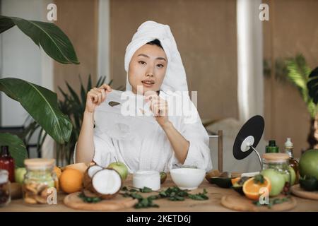 Attraktive junge asiatische Dame hält in den Händen Baumwolle Gesichtsmaske sitzt am Tisch mit verschiedenen Zutaten für hausgemachte Kosmetik. Porträt einer glücklichen Frau mit Kosmetik für die Gesichtsbehandlung. Stockfoto