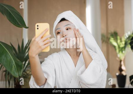 Gesunde asiatische Bloggerin, die Smartphone hält und weiße Baumwollmaske über exotischen Hintergrund aufbringt. Eine hübsche Frau, die zu Hause Hautpflegeverfahren durchführt, Selfie macht. Stockfoto