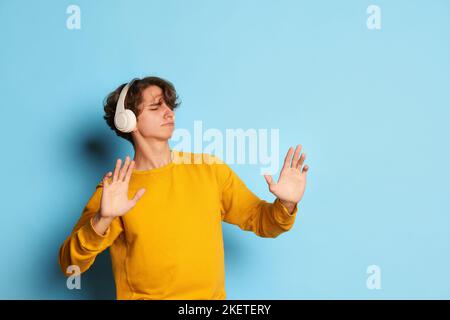 Porträt eines jungen Mannes mit lockigen Haaren, der in einem gelben Sweatshirt posiert, Musik in Kopfhörern hört und isoliert auf blauem Hintergrund tanzt. Stockfoto