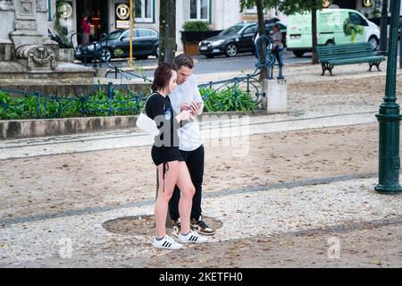 Es kommt heftiger Regen, und ein durchnässtes junges Paar nimmt mit einem Handy unter einem Baum im Miradouro de São Pedro de Alcântara in Lissabon, Portugal, Schutz. Stockfoto