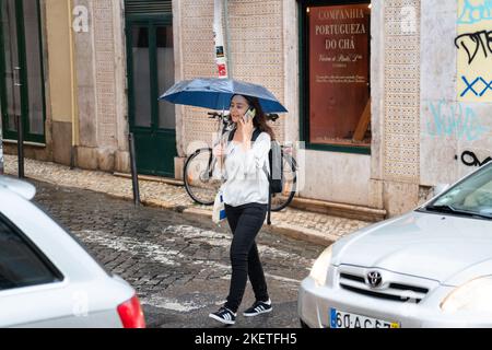 Eine Frau überquert im Sturm mit ihrem Mobiltelefon eine geschäftige Straße unter ihrem Regenschirm. Die Menschen müssen sich bei einem typischen schweren Herbstregen decken Stockfoto