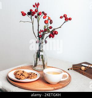 Kaffee- und Schokoladenkekse auf einem Holzserver mit Hagebuttenzweigen in Glasvase. Gemütliches Winterfrühstück. Stillleben. Stockfoto