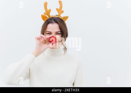 Foto einer überfröhlichen lustigen Dame in einem weihnachtlichen Hirsch-Kostüm und einem weißen Pullover mit roter Kugel und weißem Hintergrund. Stockfoto