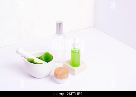 Mörtel und Stößel mit frischen grünen Ginkgo-Blättern, kosmetischen Serum- und Tonic-Flaschen, Cremeglas auf weißem Hintergrund. Natürliche handgemachte Kosmetik-Konzept. Stockfoto