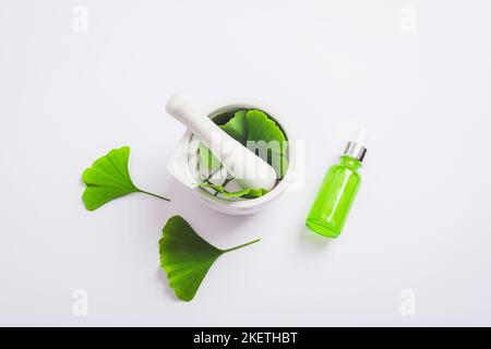 Frische grüne Ginkgo-Blätter, Mörtel und Stößel, kosmetische Serumflasche auf weißem Hintergrund. Draufsicht, flach liegend. Natürliche handgemachte Kosmetik-Konzept. Stockfoto