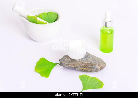 Frische grüne Ginkgo-Blätter, Mörser und Stößel, kosmetische Serumflasche und Cremeglas auf weißem Hintergrund. Natürliche handgemachte Kosmetik-Konzept. Stockfoto