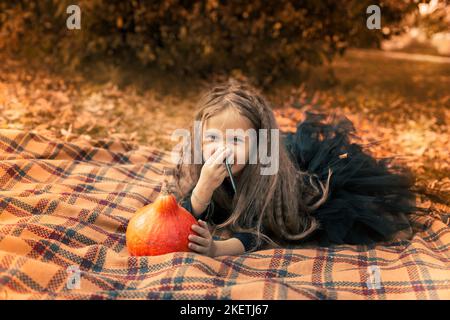 Halloween. Mädchen mit langen Haaren in schwarzem Kleid sitzt auf einer orangefarbenen Decke im Park und zieht ein Gesicht auf einen Kürbis. Stockfoto