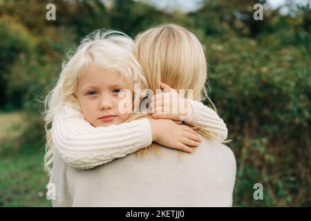 Ein kleines blondes Mädchen in den Armen ihrer Mutter umarmt ihren Hals. Stockfoto