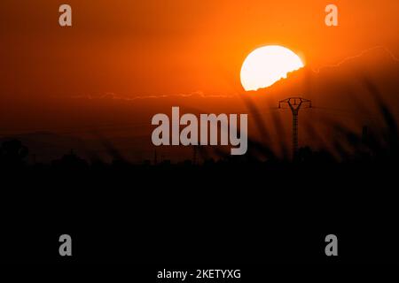 Beeindruckender Sonnenuntergang mit der Sonne, die sich hinter den Wolken über einigen Pflanzen und Hochspannungsmasten versteckt, albufera Naturpark valencia, spanien Stockfoto