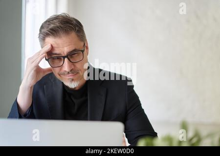 Ein unruhigen Geschäftsmann, der im Büro am Schreibtisch sitzt und mit einem Laptop arbeitet und über eine Lösung nachdenkt. Stockfoto