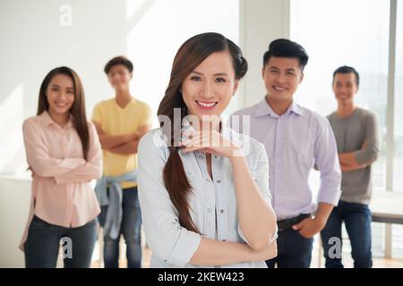Hübsche lächelnde vietnamesische junge Frau und ihre fröhlichen Mitarbeiter im Hintergrund Stockfoto