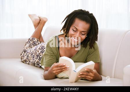 Lächelnde schwarze Frau, die auf dem Sofa liegt und ein Buch liest Stockfoto