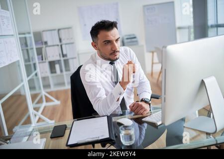 Selbstbewusster, gutaussehender Geschäftsmann, der an seinem Arbeitsplatz am Computer arbeitet Stockfoto