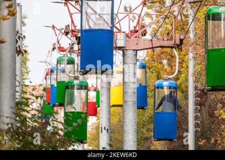 Farbenfrohe Kabinen für den Transport der Seilbahn im Herbst im Vergnügungspark der Stadt Charkiw mit goldenen Bäumen und grauem Himmel Stockfoto