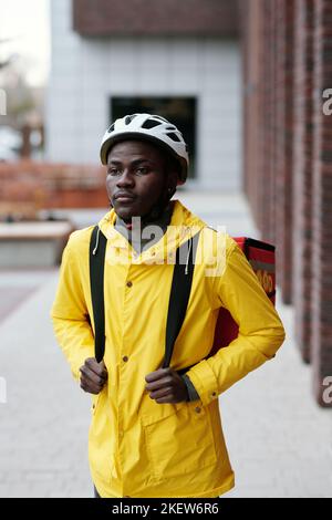 Junger ernsthafter schwarzer Mann in Schutzhelm und gelber Jacke mit Rucksack auf dem Rücken, der in der städtischen Umgebung vor der Kamera steht Stockfoto