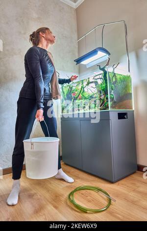 Eine Frau hält einen schweren weißen Eimer voller Wasser vor dem Aquascape des Aquariums. Starke Frau und Wasser ändern Konzept. Stockfoto