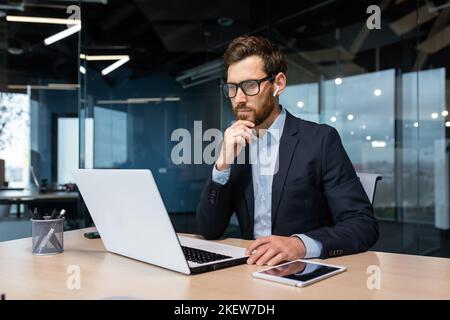Ein leitender Geschäftsmann mit Brille und Bart denkt über eine Entscheidung nach, ein Mann arbeitet im Büro und benutzt einen Laptop bei der Arbeit, ein reifer Investor in einem Geschäftsanzug sitzt am Tisch Stockfoto