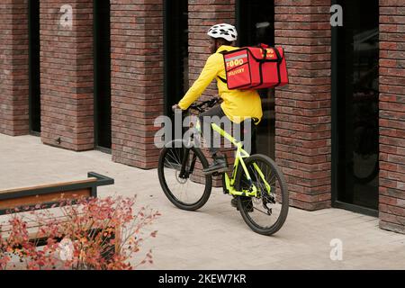 Rückansicht eines jungen Kuriers in Jeans, Jacke und Helm, der mit dem Fahrrad entlang der Backsteinmauer des modernen Gebäudes fährt, während er den Kunden Lebensmittel liefert Stockfoto