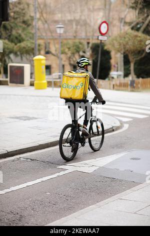 Spanien, Salamanca - 3. April 2022: Lieferung von Lebensmitteln und Lebensmitteln. Der Fahrradkurier mit gelbem Thermo-Rucksack, die Firma Glovo, liefert das nötige Stockfoto