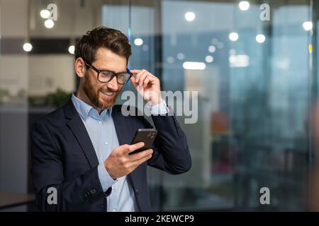 Erfolgreicher Finanzinvestor arbeitet im Büro bei der Arbeit, Geschäftsmann im Geschäftsanzug nutzt Telefon in der Nähe des Fensters, Mann lächelt und liest gute Nachrichten online vom Smartphone. Stockfoto