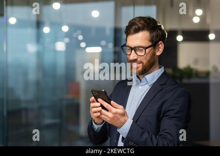 Erfolgreicher Finanzinvestor arbeitet im Büro bei der Arbeit, Geschäftsmann im Geschäftsanzug nutzt Telefon in der Nähe des Fensters, Mann lächelt und liest gute Nachrichten online vom Smartphone. Stockfoto