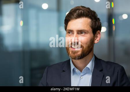 Nahaufnahme eines glücklichen und lächelnden, reifen Geschäftsmannes im modernen Büro, der zur Seite schaut, und eines Mannes im Geschäftsanzug bei der Arbeit. Stockfoto