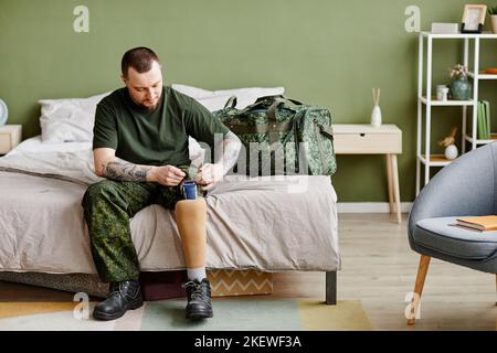Porträt eines Militärveteranen in voller Länge mit prothetischem Bein, das im Schlafzimmer eine Armeeuniform aufsetzt und Platz kopiert Stockfoto