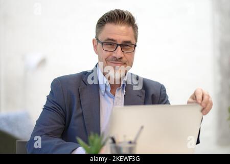 Ein Geschäftsmann mittleren Alters, der am Schreibtisch mit einem Laptop arbeitet. Mann im Business Casual sitzt im Büro. Stockfoto