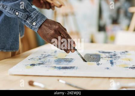 Selektiver Fokus auf die Hand des jungen schwarzen Mannes Malerei auf weißer Leinwand mit Acrylfarben, während sie am Tisch im Atelier der Künste stehen Stockfoto