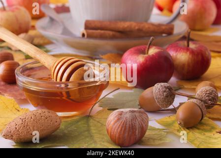 Weißer Tisch mit Herbstschmuck, einer Tasse Tee, Zimtstücken, einer Glasschale mit Honig, kleinen Äpfeln, Nüssen, bunten Baumblättern und Eicheln. Niedrig Stockfoto