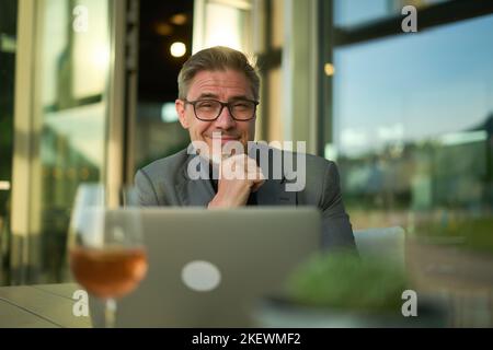 Glücklicher Geschäftsmann mit Laptop-Computer im Restaurant. Mann mittleren Alters, der am Tisch in der Hotellobby oder in der Business Lounge im Büro sitzt, arbeitet und lächelt. Stockfoto
