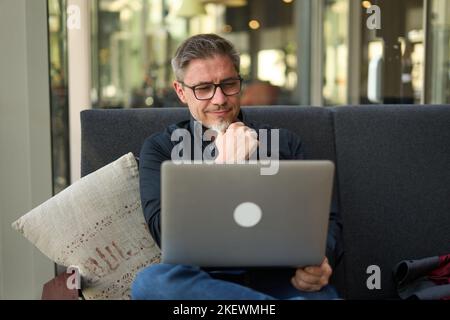 Geschäftsporträt - Geschäftsmann sitzt auf dem Sofa und arbeitet mit einem Laptop im Büro, in der Lobby oder im öffentlichen Raum, glücklicher Mann mittleren Alters lächelt und denkt nach. Stockfoto