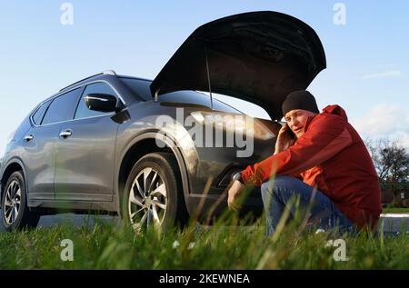 Frustrierter männlicher Fahrer sitzt mit offener Motorhaube in der Nähe eines kaputten Autos und ruft am Telefon an. Transportkonzept. Stockfoto