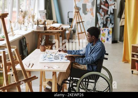 Junge zeitgenössische Künstlerin im Rollstuhl arbeitet über neue Malerei, während sie im Atelier der Künste sitzt, Farben mischt und auf Leinwand aufsetzt Stockfoto