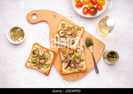 Holzbrett mit leckeren Toasts mit Frischkäse, Pilzen und Pesto-Sauce auf hellem Hintergrund Stockfoto