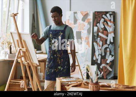 Junger ernsthafter afroamerikanischer Mann in Jeanschürze, der auf Staffelei vor der Leinwand steht und auf der Freizeit neues Gemälde kreiert Stockfoto