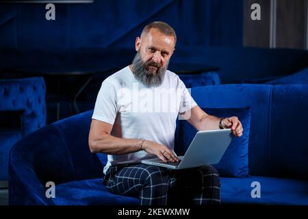 Erwachsener männlicher Geschäftsmann, Lehrer, Mentor, der an einem neuen Projekt arbeitet. Sitzt an einem großen Fenster auf dem Tisch. Er schaut auf den Laptop-Bildschirm. Stockfoto