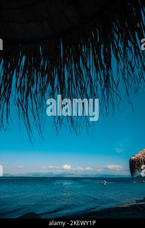 Blick Auf Das Ionische Meer, In Dem Urlauber Schwimmen. Im Vordergrund Ist Silhouette Des Strandschirms Aus Palmblättern, Griechenland, Moraitika, Korfu. Stockfoto