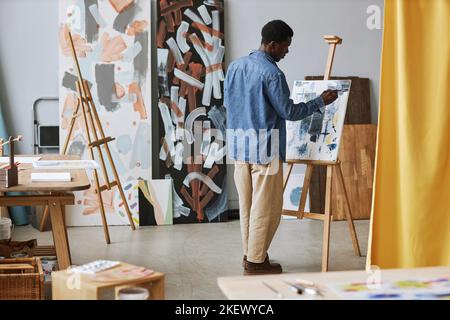 Zurück von jungen schwarzen Mann in Casualwear arbeiten über neue Malerei, während vor Staffelei mit Leinwand bei der Lektion in der Schule der Künste stehen Stockfoto