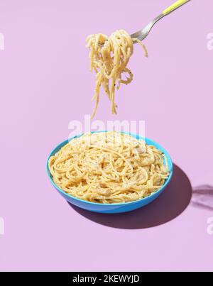 Pasta Cacio e Pepe in einer blauen Schale isoliert auf einem violetten Hintergrund. Spaghetti mit einer Gabel aus einer Schüssel in hellem Licht gegen einen bunten Backgro nehmen Stockfoto