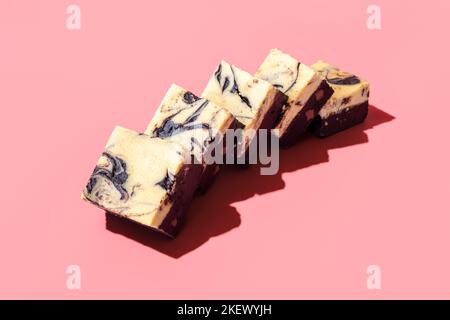 Hausgemachter, geschichteter Kuchen mit einer Schicht Schokolade und einem Käsekuchen. Brownie-Scheiben mit Käsekuchen in hellem Licht auf einem rosafarbenen Tisch Stockfoto