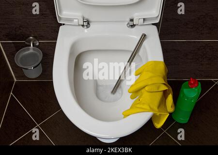 Toilettenschüssel mit Toilettenbürste, gelben Gummihandschuhen und einem Reinigungs- und Desinfektionsmittel auf dem Hintergrund dunkler Keramikfliesen im Toilettenraum Stockfoto