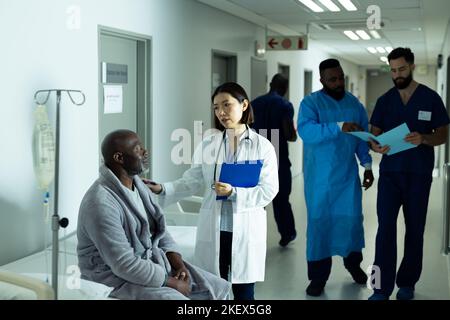 Asiatische Ärztin tröstet ranghohen afroamerikanischen männlichen Patienten im Krankenhauskorridor Stockfoto