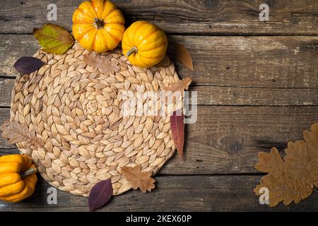 Gemütlicher Hygge-Tisch. Draufsicht auf Weidenplatz-Matte, Kürbisse, Herbstblätter, Kerze auf dunklem Tisch. Herbsttischeinstellung. Festliches Erntedankfest im Hintergrund. Stockfoto