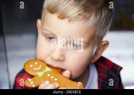 Nettes kaukasisches Kind isst Lebkuchen und macht einen lustigen Gesichtsausdruck. Weihnachtliches Essen. Stimmungsvolles, ästhetisches Weihnachtshaus für Silvester. Kopf Hoch Stockfoto