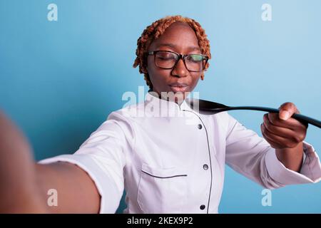 afroamerikanische Köchin bläst Löffel mit heißer Suppe. Professionelle Köchin mit geschlossenen Augen trägt Küchenuniform in Studio Shot isoliert in blau, Kochkonzept. Stockfoto