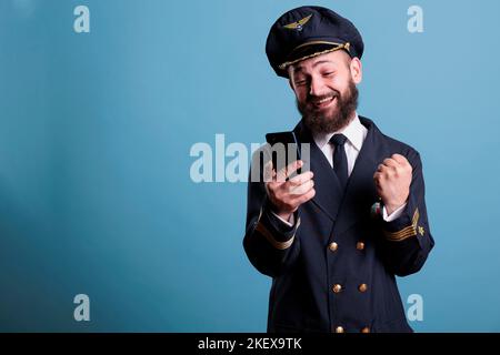 Glücklich aufgeregt Flugzeug Flieger in Uniform hält Smartphone, so dass Sieger Geste, geballte Faust. Pilot mit fröhlichem Gesichtsausdruck, der auf den Bildschirm des Mobiltelefons schaut und gute Nachrichten liest Stockfoto