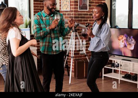 Fröhliches afroamerikanisches Paar, glücklich tanzend, Wein trinkend, Alkohol begleitet von kaukasischer junger erwachsener Freundin. Partner genießen Freundschaft, Partnerschaft, soziales Leben. Stockfoto