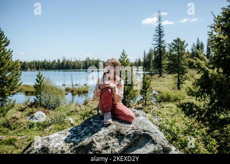 Junges Mädchen, das draußen auf einem Felsen neben dem See sitzt Stockfoto