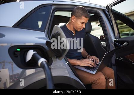 Ein Mann mit Laptop sitzt im Elektroauto und wird an der Station aufgeladen Stockfoto